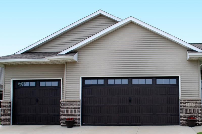 Dark brown garage doors with window inserts on a tan vinyl-sided home with brick columns