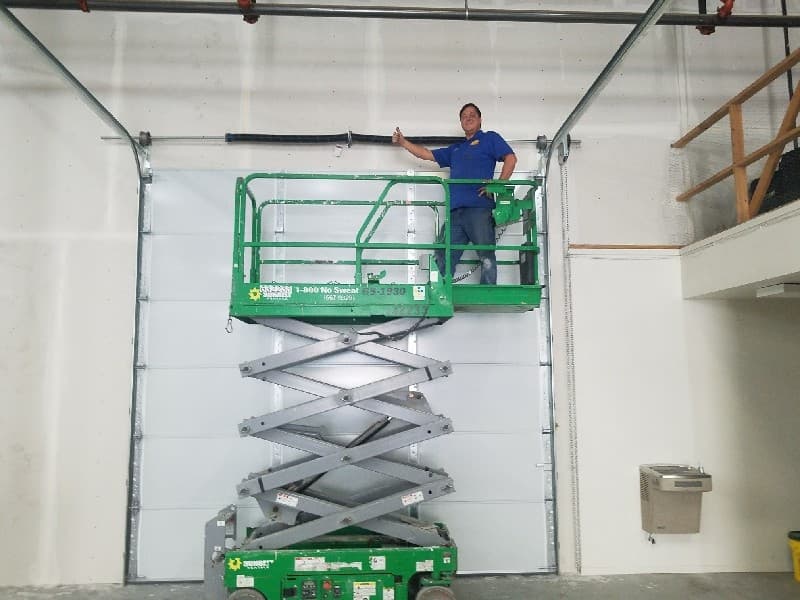 Technician on a scissor lift beside a tall commercial sectional door and its torsion spring
