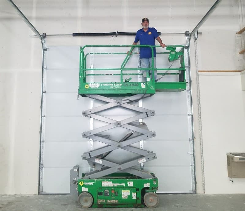 Technician on a scissor lift inspecting the torsion spring above a large white commercial garage door