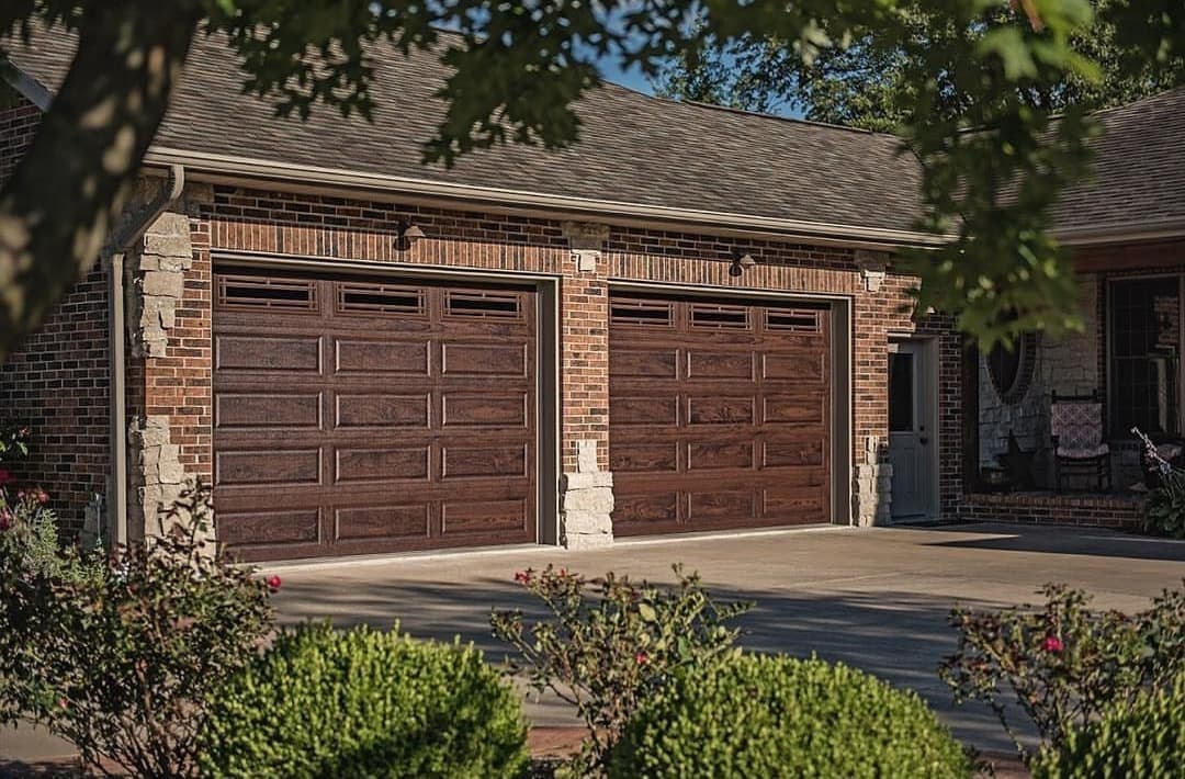 Two brown wood-grain garage doors with decorative windows on a red brick home with stone accents