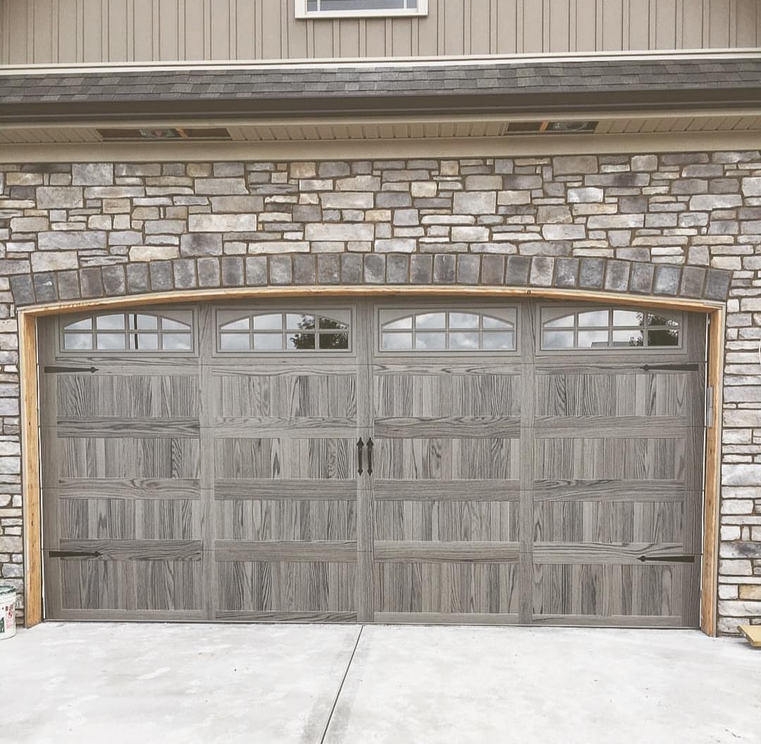 Gray wood-look carriage-style garage door with arched windows and black hinges under a stone arch